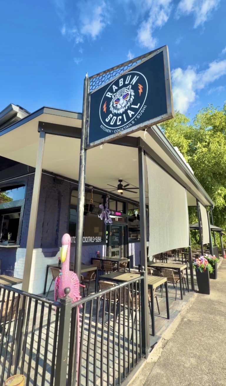 Exterior view of Rabun Social restaurant with a large black sign showing a tiger wearing sunglasses and the text ‘Kitchen Cocktails Sushi,’ outdoor seating under a covered patio.