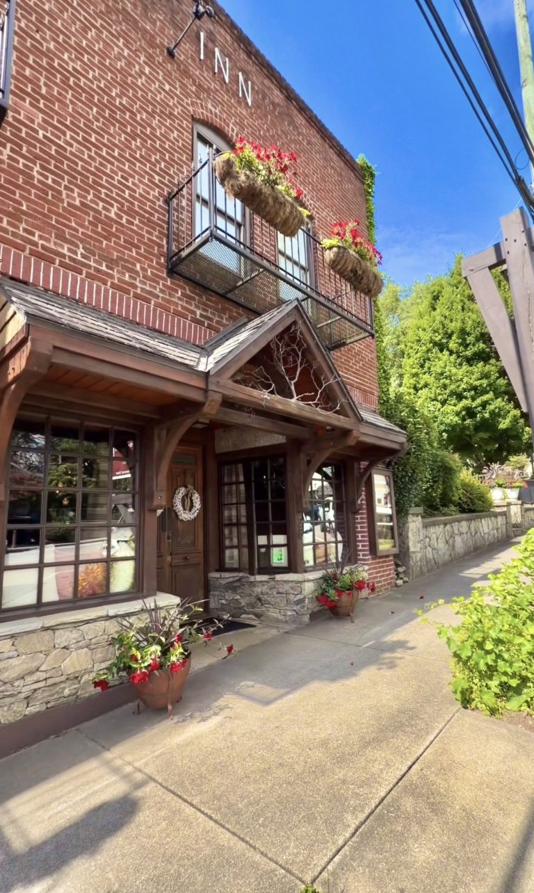 Brick inn with a metal balcony planter filled with red flowers on the second floor. A wooden storefront with a wreath sits below.