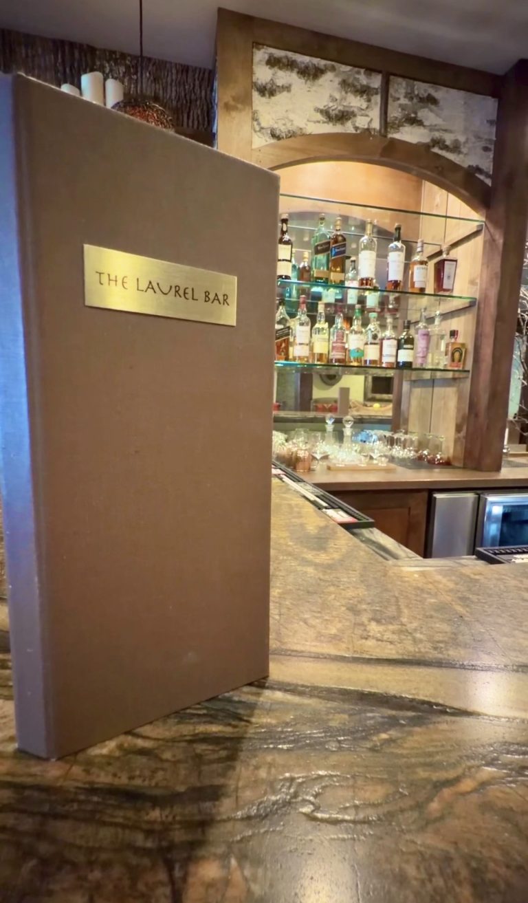 Brown divider with a brass plaque reading 'The Laurel Bar' next to a backlit liquor shelf and glassware behind.