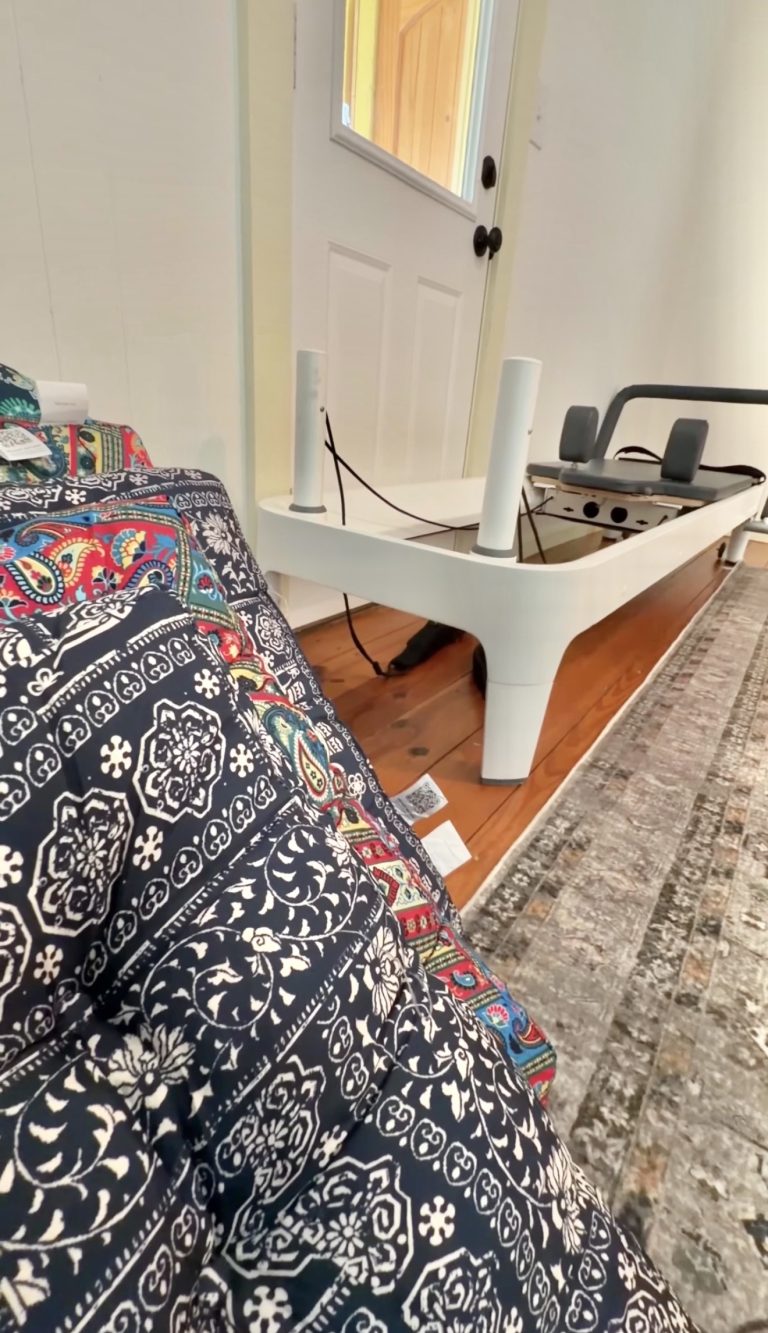 White Pilates reformer machine in a bright room, with a colorful patterned quilt in the foreground and a closed white door in the background.