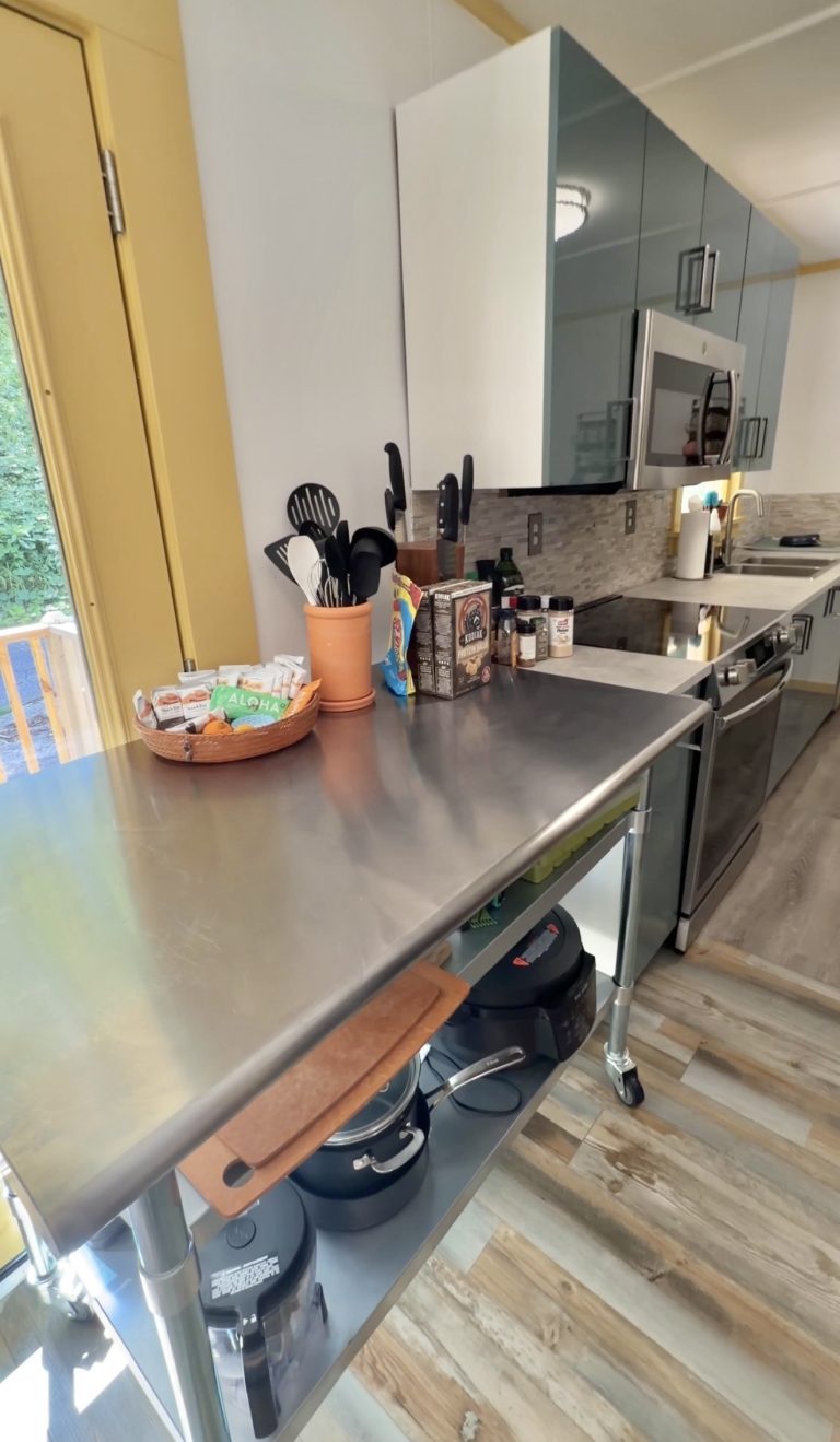 Stainless steel kitchen island with utensil holder, spices, and cookware beside gray upper cabinets and stainless appliances.